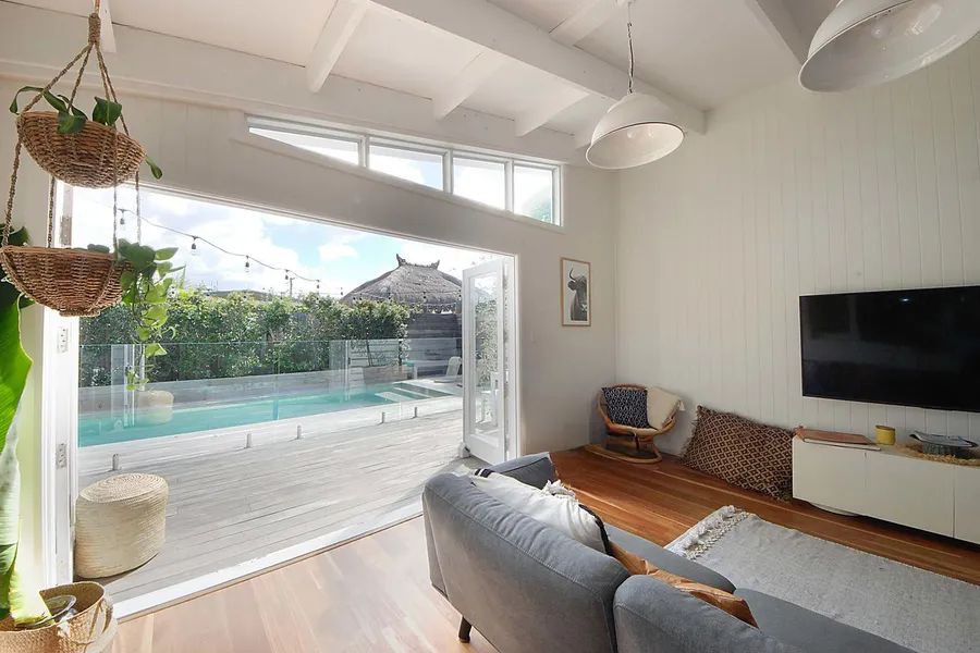 Interior of a renovated living room with high clerestory windows above wide, open bifold doors leading out to a deck and swimming pool