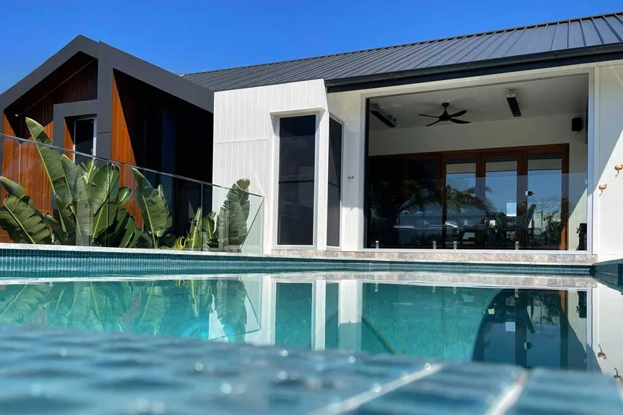 View of the house facade from the infinity pool, combining natural timber, white cladding, and dark metallic look panels