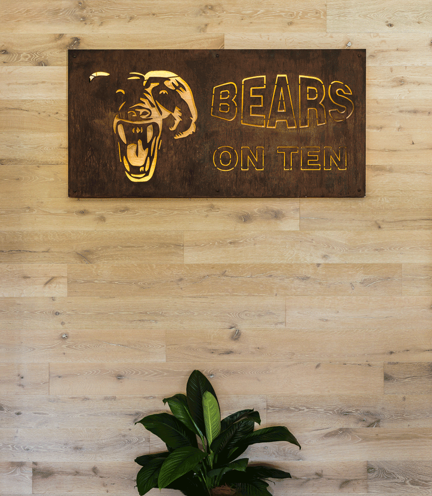 Elegant signage of a sporting club etched onto a corten steel sign affixed to a rustic oak timber-clad wall, accompanied by a lush potted plant.